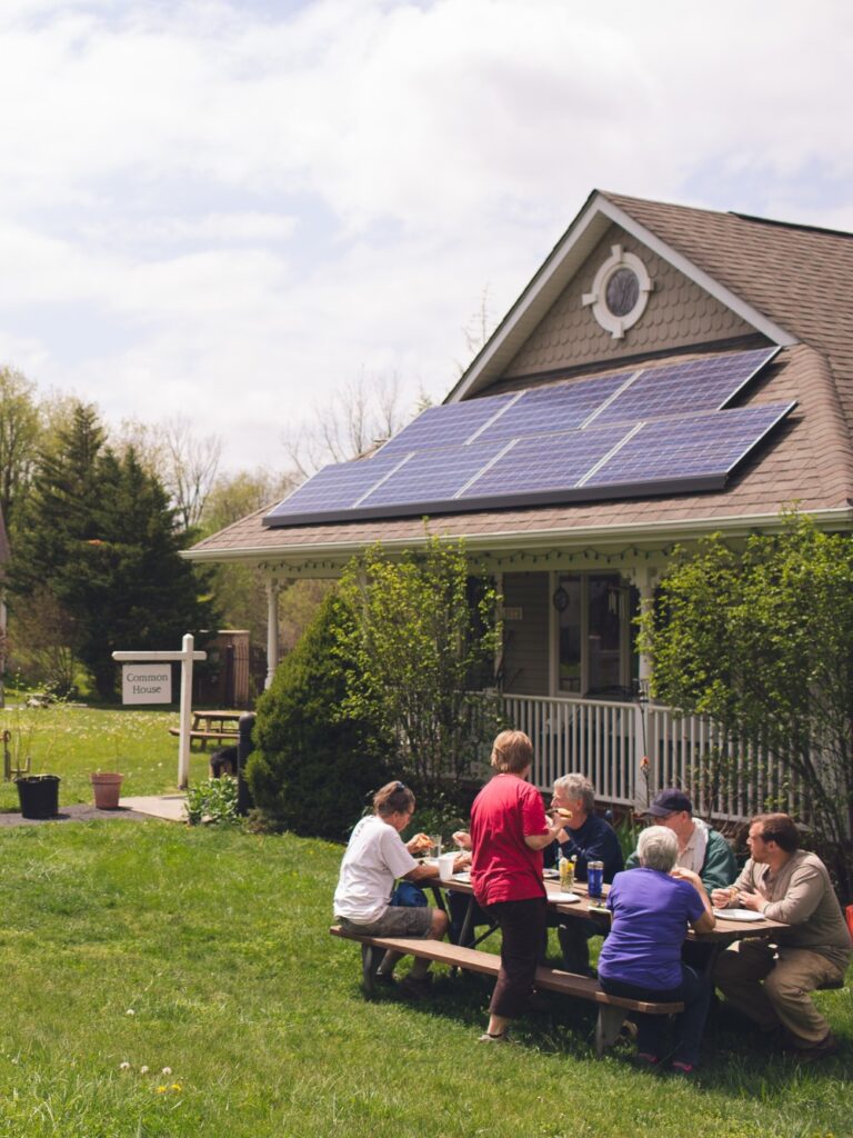 Solar panels on the roof of a duplex with a "Common House" sign in front. People of all ages are sitting at a picnic bench out front.