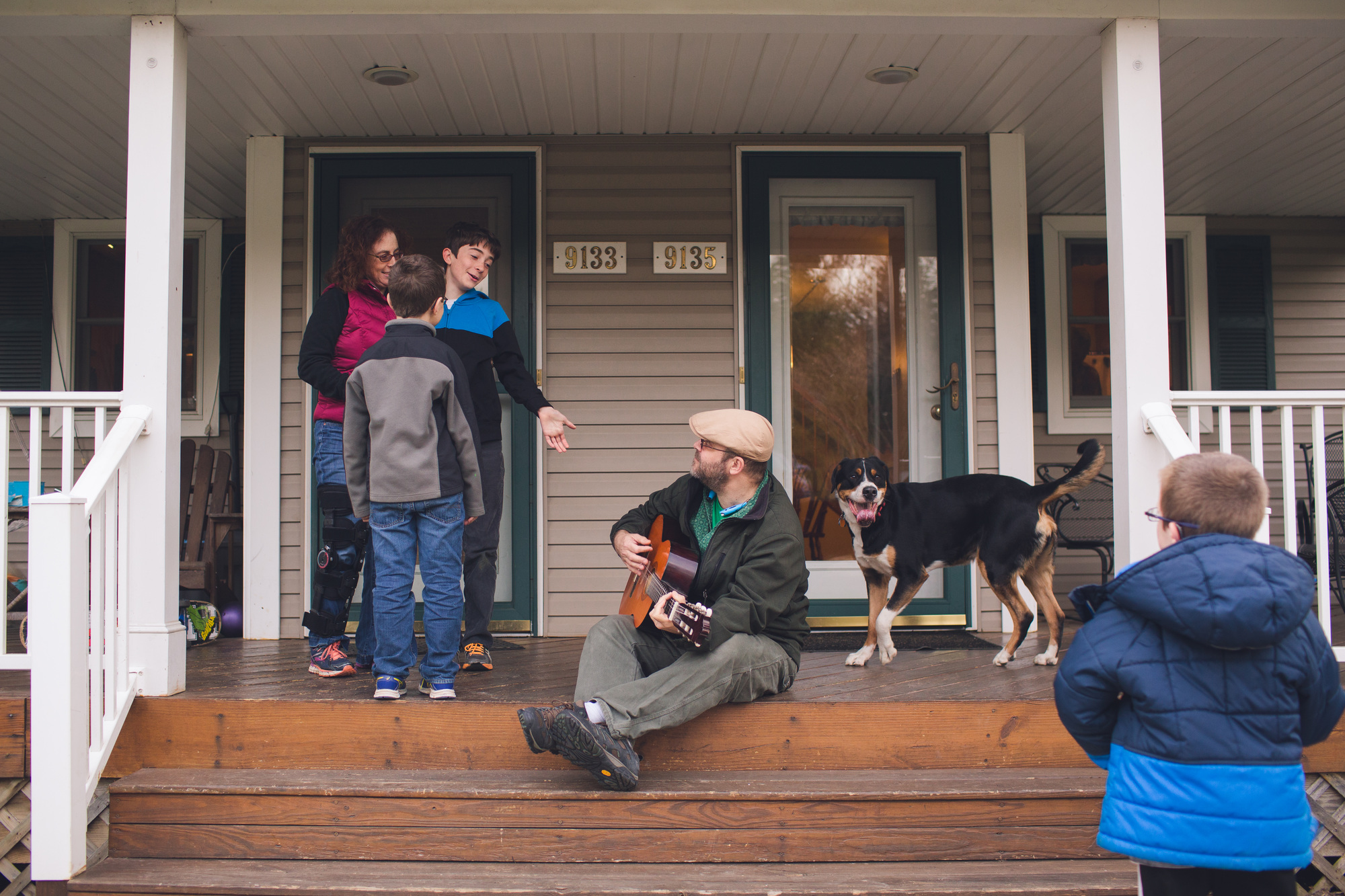 Man with guitar sitting on porch, children standing nearby and a dog looking happy.